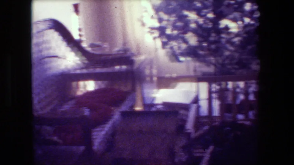 1981 Hollywood home-movie frame showing a wicker chair, window light, and a Christmas tree in a private interior with purple-shifted 8mm color.