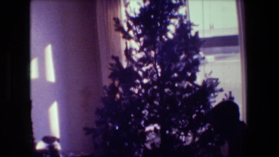 1981 Hollywood home-movie frame showing a Christmas tree near a curtained window, with daylight patches falling across the wall in a private interior.