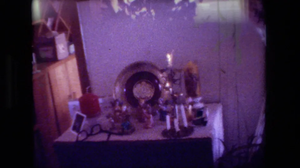 1981 Hollywood home-movie frame showing a holiday tabletop with a lit candle, small decorations, framed items, and surrounding furniture in a private interior.