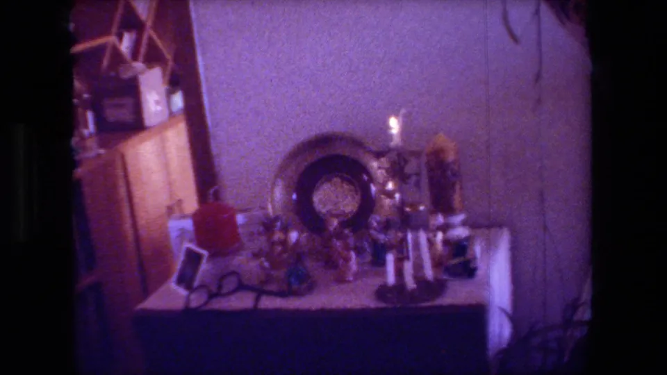 Hollywood, California home-movie frame from 1981 showing a dim private living room table arranged with candles, a decorative plate, small ornaments, and a red container.