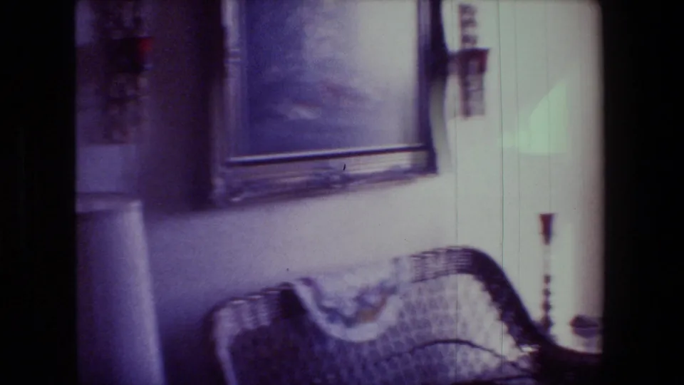 1981 Hollywood home-movie frame showing a wicker chair beneath a framed wall picture, with candles and nearby living room decor in a private interior.