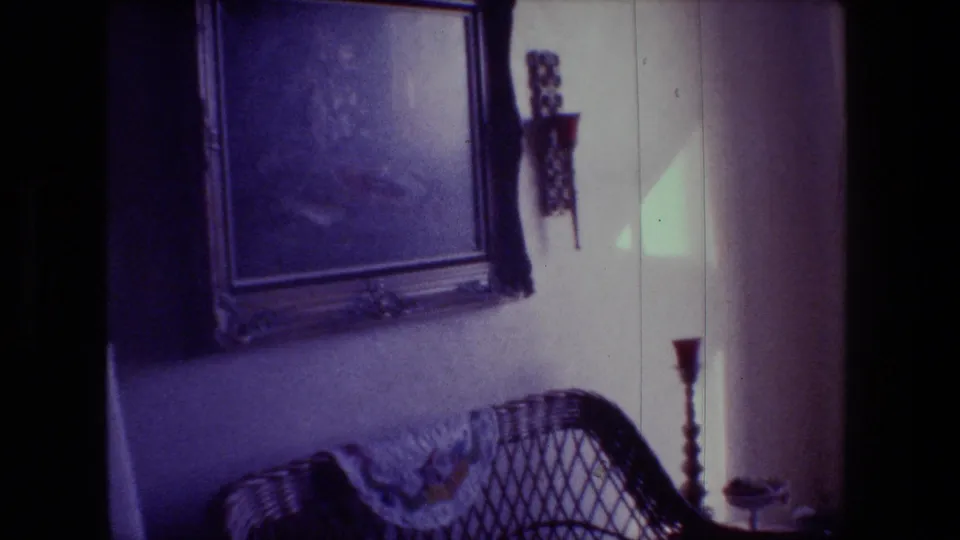 1981 Hollywood, California home-movie frame showing a wicker chair, framed wall art, wall sconce, and candlestick holder in a softly lit living room.