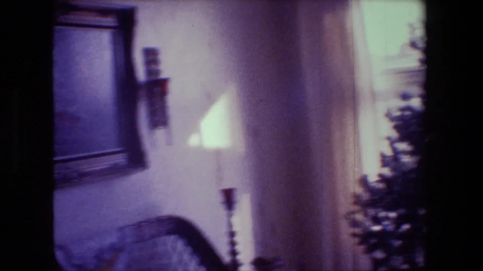 1981 Hollywood, California home-movie frame with framed wall art, candle fixtures, bright window light, and the edge of a Christmas tree inside a private living room.