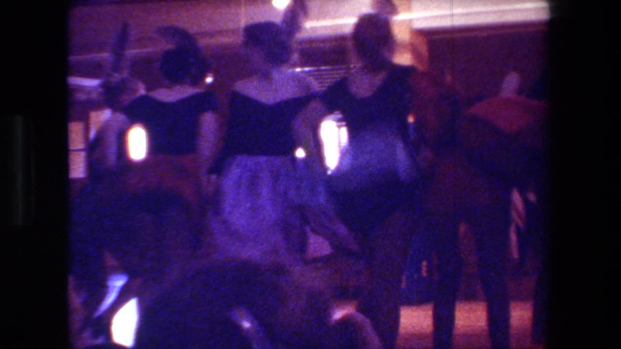 In 1981 Hollywood archival Super 8 footage, adult female dancers in black tops and full skirts move in formation under warm lights in a dark club or theater interior.