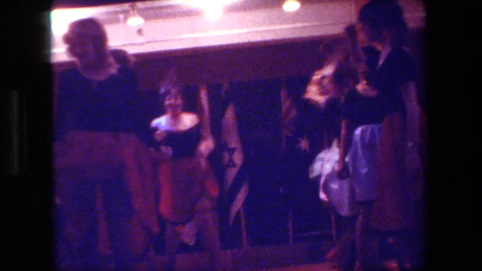 Adult dancers in a dark Hollywood interior perform a cancan-style routine, with one figure near the camera and others visible against the back wall.