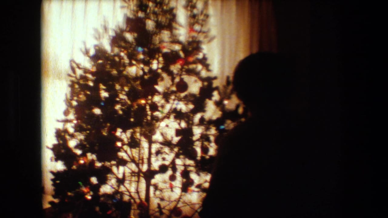 In this 1981 Lansing, Michigan home-movie frame, a silhouetted person stands near a lighted Christmas tree set against sheer curtains and warm indoor light.