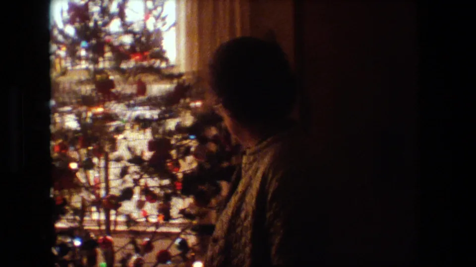A rear-view silhouette of a person faces a decorated and lighted Christmas tree near a curtained window in the supplied 1981 Lansing home-movie setting.