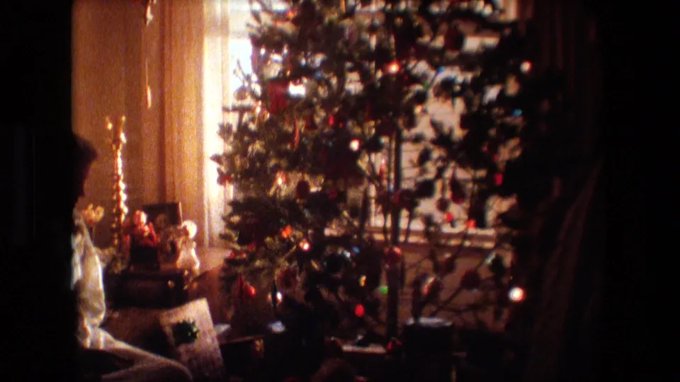 A decorated Christmas tree with colored ornaments stands near a window in a warm private interior, with small holiday items visible near the base and side table area.
