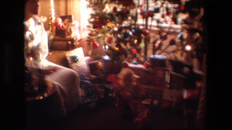 The frame shows the Christmas tree area with ornaments, tinsel, presents, and nearby furniture in the same warm Lansing home interior.