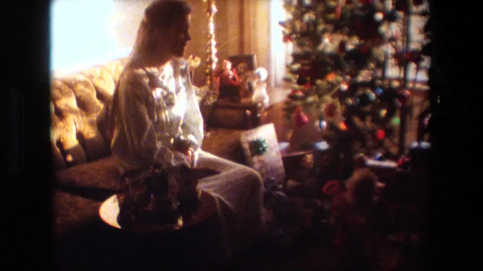 A woman in a light-colored nightgown sits on a couch beside the decorated Christmas tree and wrapped gifts in this 1981 Lansing home-movie frame.