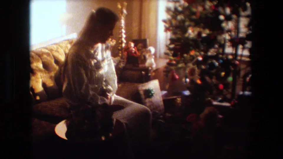 The woman remains seated in profile near the tree, with ornaments, tinsel, gifts, and warm window light visible in the private Lansing living room.