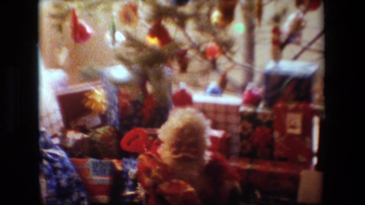 1981 Lansing, Michigan home-movie frame showing a decorated Christmas tree, wrapped presents, and holiday figurines in a warm living room interior.