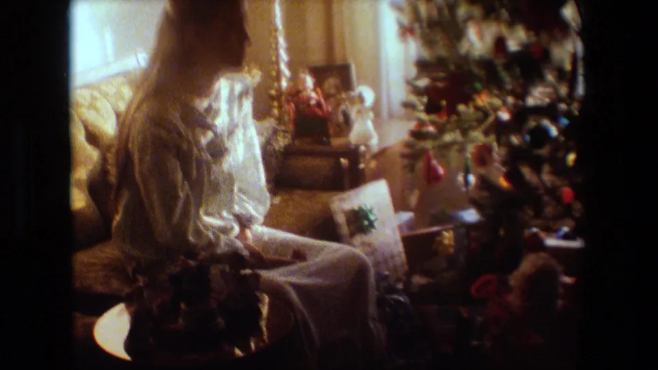 1981 Lansing, Michigan home-movie frame showing a woman in a light nightgown or robe seated beside a decorated Christmas tree and wrapped presents.