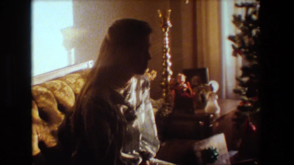 1981 Lansing, Michigan home-movie frame showing a woman in profile near a tufted sofa, candlesticks, holiday figurines, and the edge of a Christmas tree.