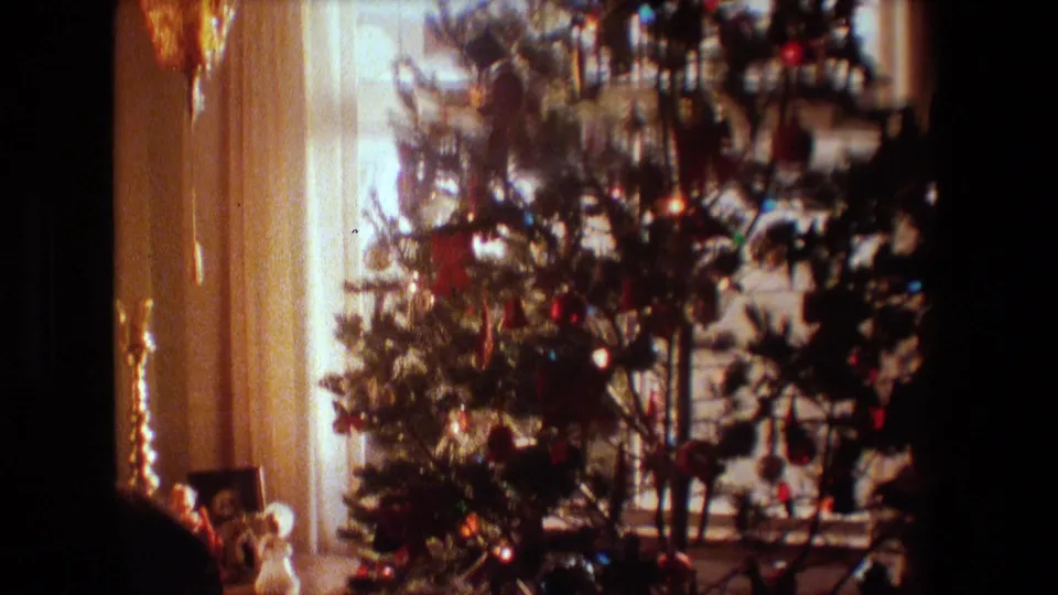 A decorated Christmas tree with colored ornaments and small lights stands near a curtained window in a dim Lansing, Michigan home interior from the 1981 8mm footage.
