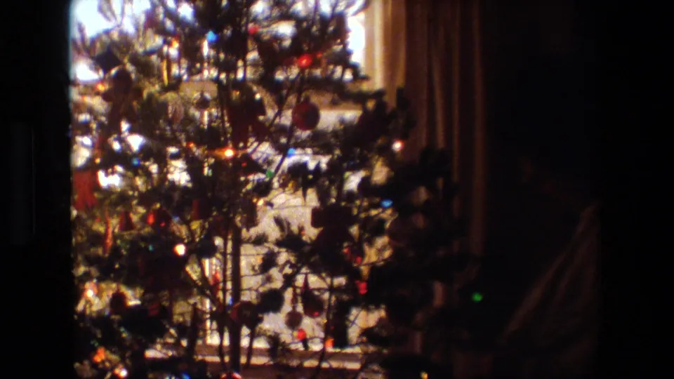 A closer 8mm view shows branches, colored ornaments, and glowing lights on the Christmas tree near the window inside the Lansing home.