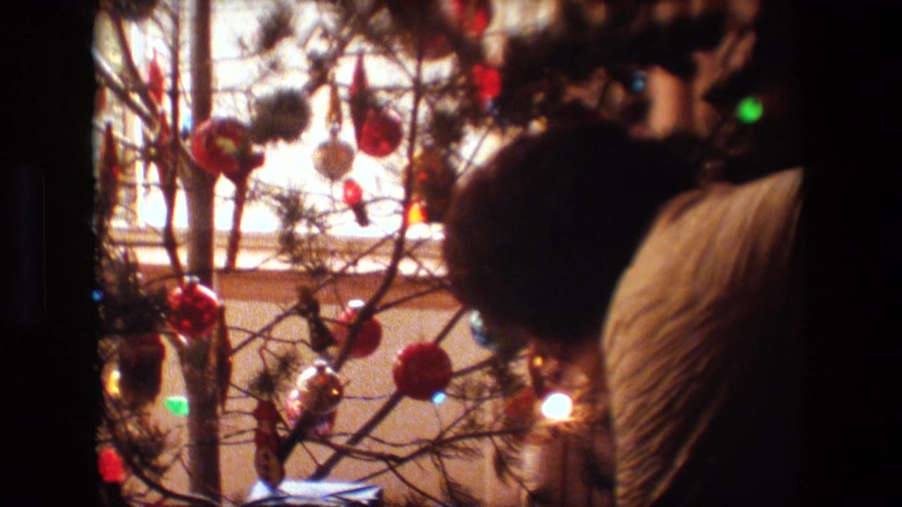 Lansing, Michigan 1981 home-movie frame showing an adult in a quilted jacket leaning near a small Christmas tree with red ornaments, lights, and wrapped gifts below.