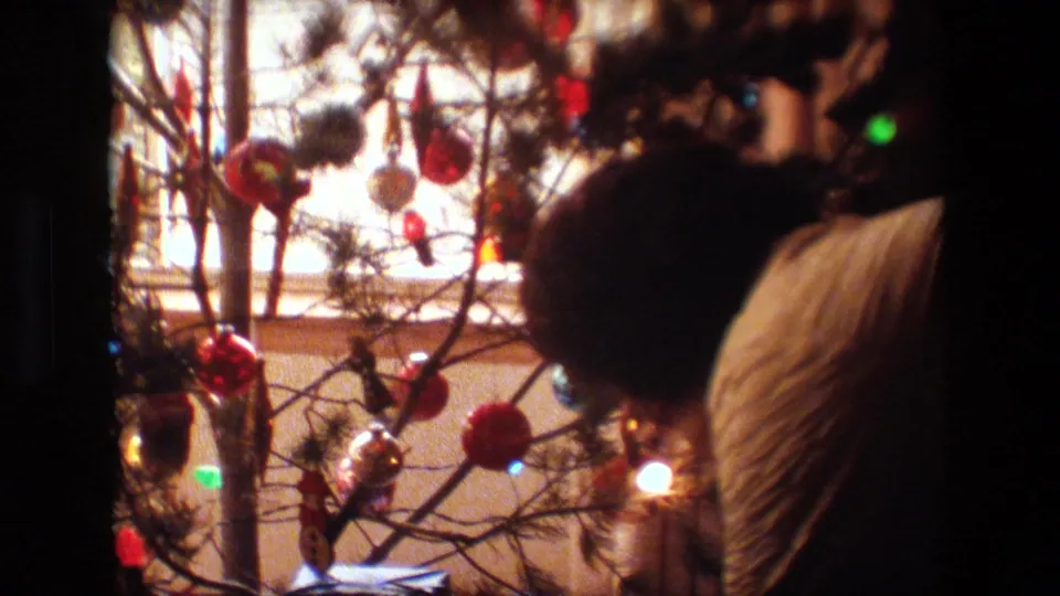 Lansing, Michigan 1981 home-movie frame showing an adult in a quilted jacket leaning near a small Christmas tree with red ornaments, lights, and wrapped gifts below.
