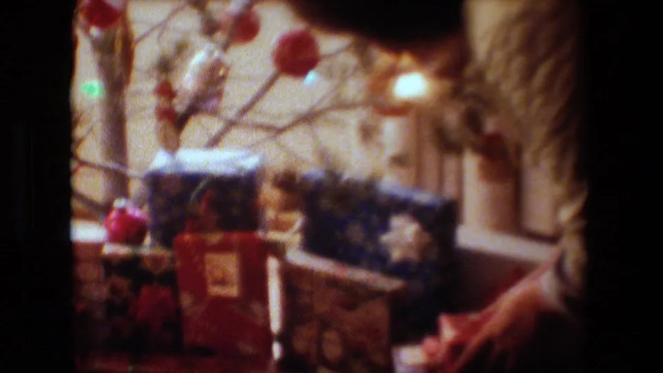 Lansing, Michigan 1981 frame with wrapped gift boxes beneath a small Christmas tree, ornaments hanging above, and part of an adult in a quilted jacket at the side.