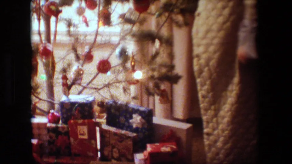 A soft 8mm Lansing 1981 frame shows a small Christmas tree with red ornaments, lights, and wrapped gifts, with an adult figure partly visible at the right edge.