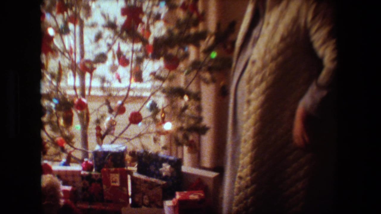 In this 1981 Lansing, Michigan home-movie frame, a woman in a quilted robe stands partly in shadow beside a decorated Christmas tree with lights and wrapped gifts near a window.