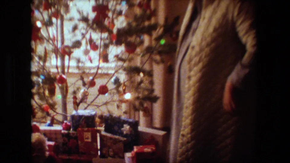 In this 1981 Lansing, Michigan home-movie frame, a woman in a quilted robe stands partly in shadow beside a decorated Christmas tree with lights and wrapped gifts near a window.