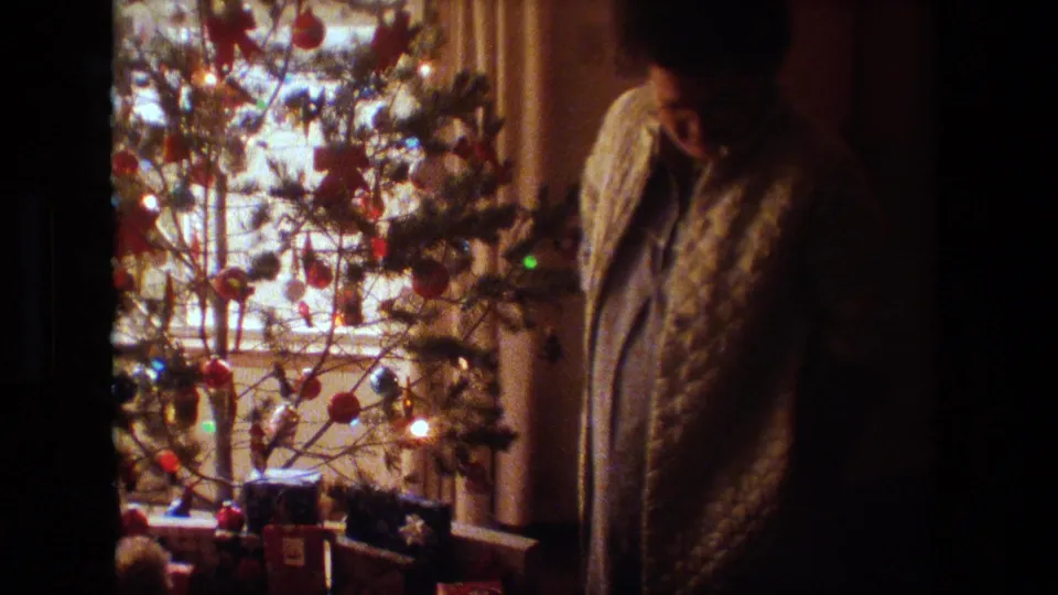 A woman in a quilted robe stands in profile beside a softly lit Christmas tree in a private Lansing home interior, with curtains and wrapped presents visible nearby.