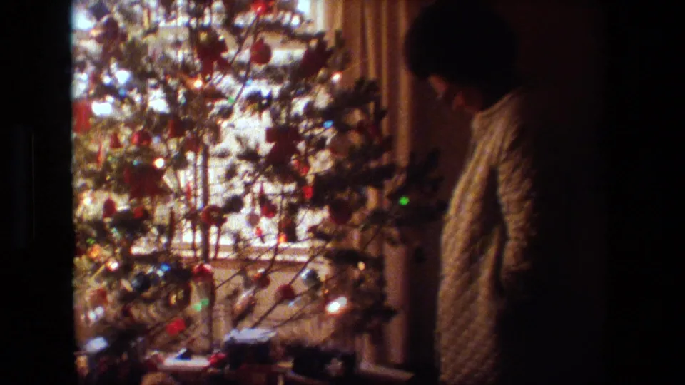 The woman appears to look downward beside the Christmas tree, with bright window light, ornaments, and soft 8mm grain shaping the private holiday scene.