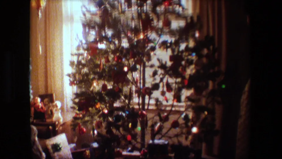 1981 Lansing, Michigan home-movie frame showing a decorated indoor Christmas tree in front of bright sheer-curtained windows, with small objects near the base.