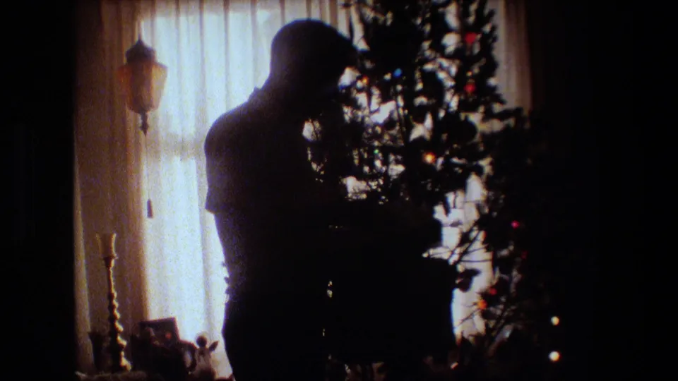 1981 Lansing, Michigan home-movie frame showing an adult man in silhouette adjusting a Christmas tree near bright sheer-curtained windows inside a private room.