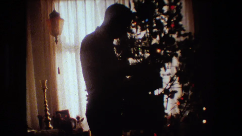 1981 Lansing, Michigan frame of a silhouetted adult man working near the branches of a decorated Christmas tree, with sheer curtains and household objects behind him.