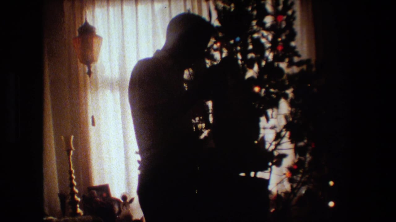In 1981 Lansing, Michigan home-movie footage, a silhouetted man stands near a Christmas tree in a dim living room with a bright curtained window behind him.