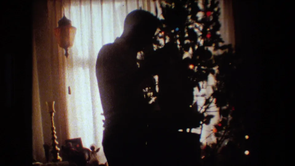 In 1981 Lansing, Michigan home-movie footage, a silhouetted man stands near a Christmas tree in a dim living room with a bright curtained window behind him.