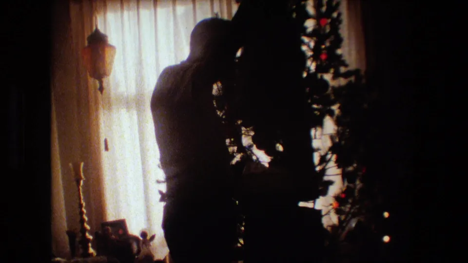 The man remains in silhouette as he leans toward the Christmas tree, with the curtained window and living room objects visible in the backlight.