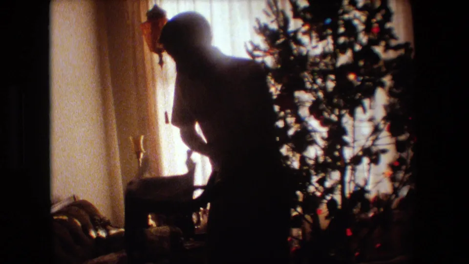 The man bends beside the Christmas tree near wrapped items or decor, with the bright curtained window and household furnishings framing the 1981 home-movie scene.