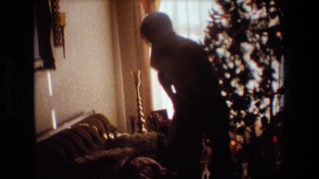 In a 1981 Lansing, Michigan home-movie frame, a silhouetted adult man stands beside a Christmas tree in a backlit living room with curtains and upholstered furniture.