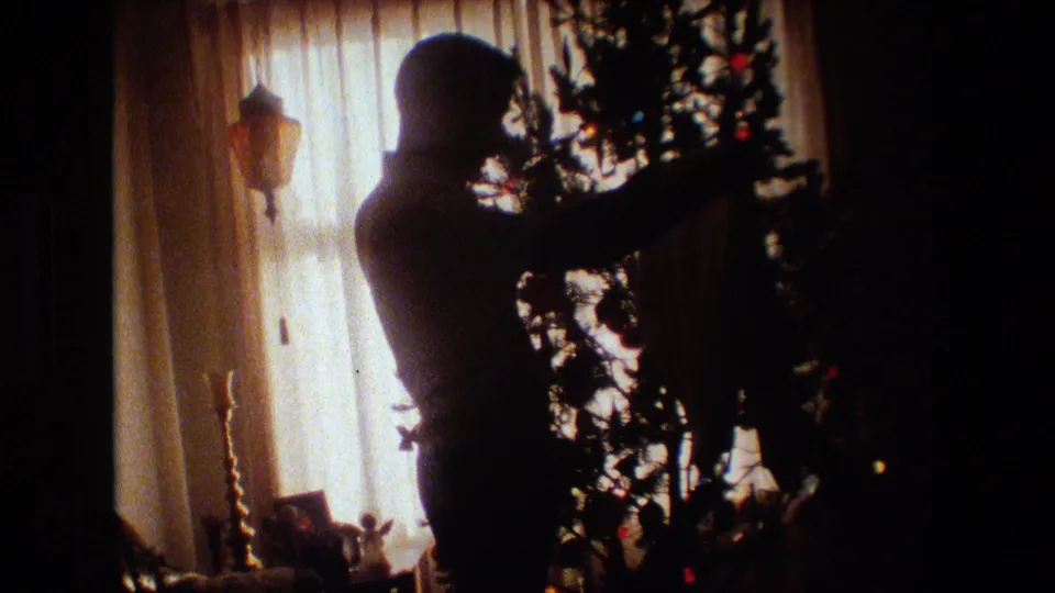 The silhouetted figure of an adult man extends an arm into a Christmas tree inside a dim 1981 Lansing, Michigan living room with curtains and holiday decorations.