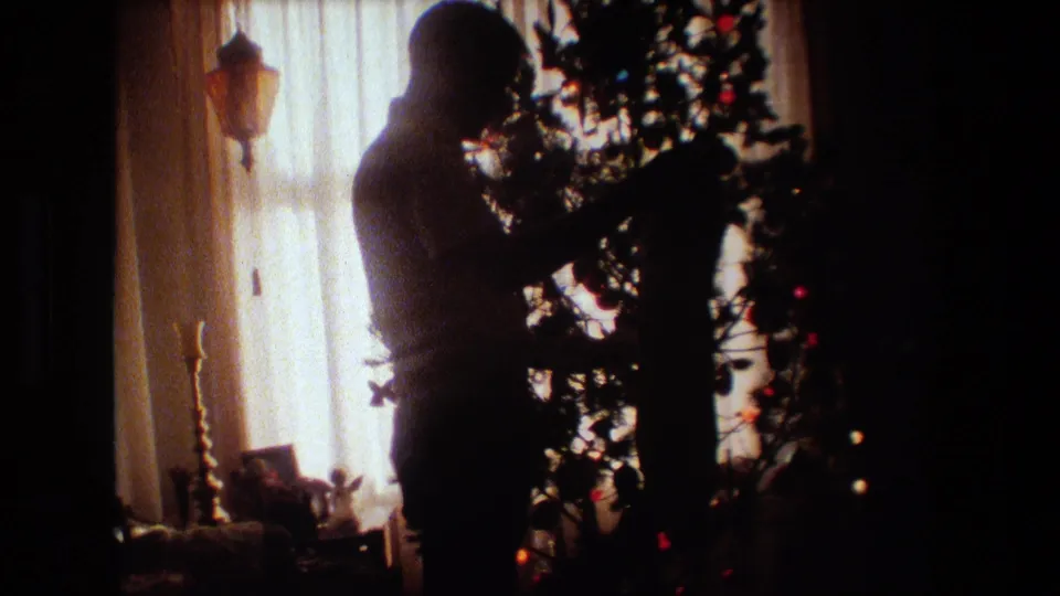 In a 1981 Lansing, Michigan home interior, a silhouetted adult man stands near a decorated Christmas tree backlit by warm window light.