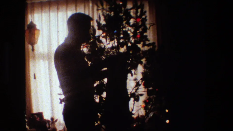 A silhouetted adult man stands close to the Christmas tree, with ornaments and small lights visible against the bright curtained window.