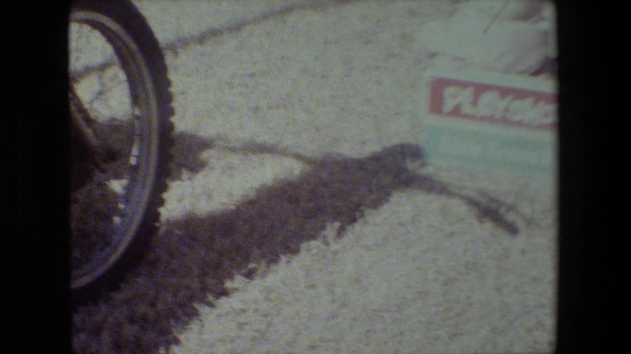 In 1981 Lansing, Michigan home-movie footage, a yellow motorcycle wheel appears at the edge of frame near a small PlaySkool box wagon on bright gravel.