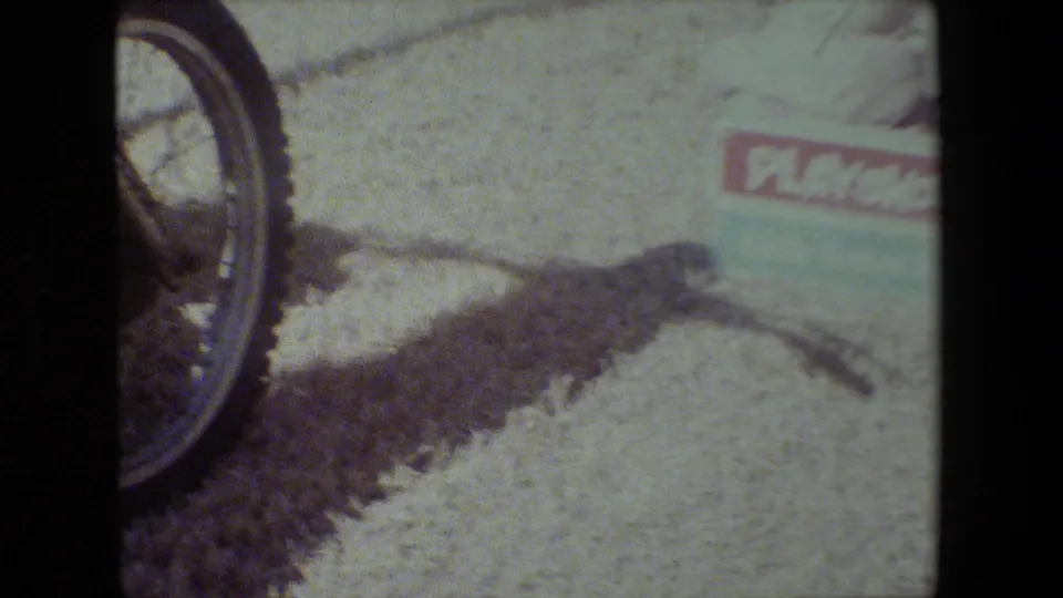 In 1981 Lansing, Michigan home-movie footage, a yellow motorcycle wheel appears at the edge of frame near a small PlaySkool box wagon on bright gravel.