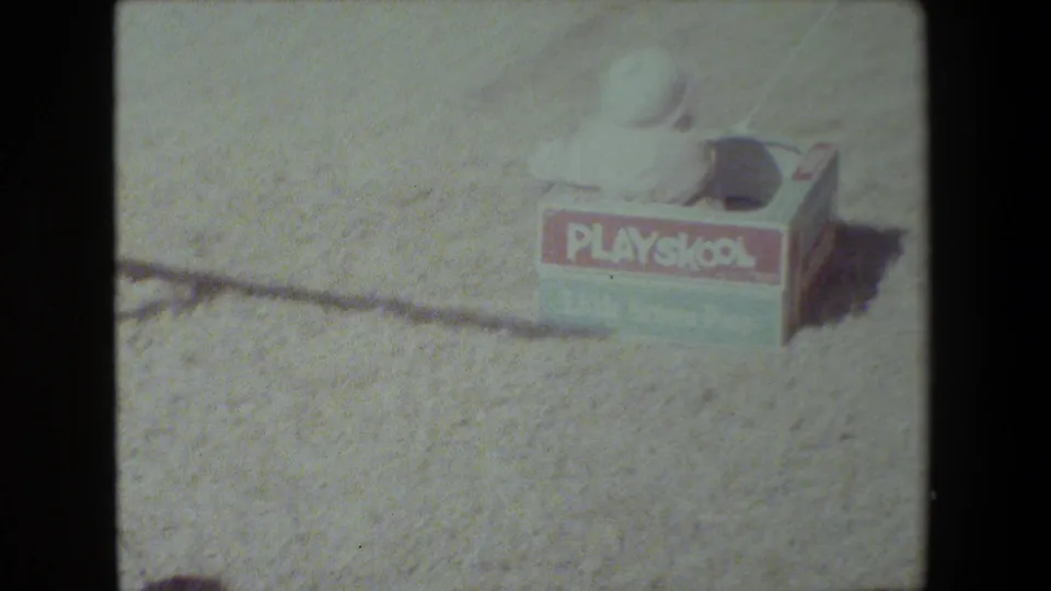 A child wearing a light-colored hat sits in a PlaySkool box wagon on sunlit gravel in the Lansing, Michigan home-movie scene.
