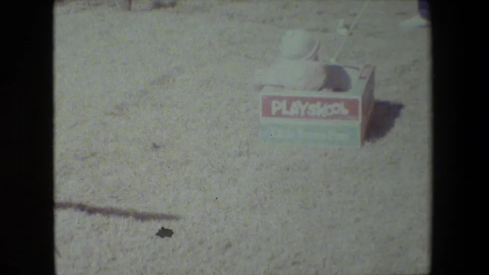 The color-faded 8mm frame shows the small PlaySkool wagon and child in the bright gravel yard, with details softened by exposure and film grain.