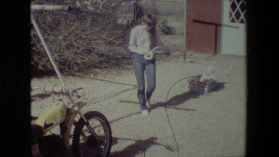 An adult in a light shirt and jeans pulls the child’s PlaySkool wagon by rope across a sunlit Lansing yard while a yellow motorcycle sits nearby.