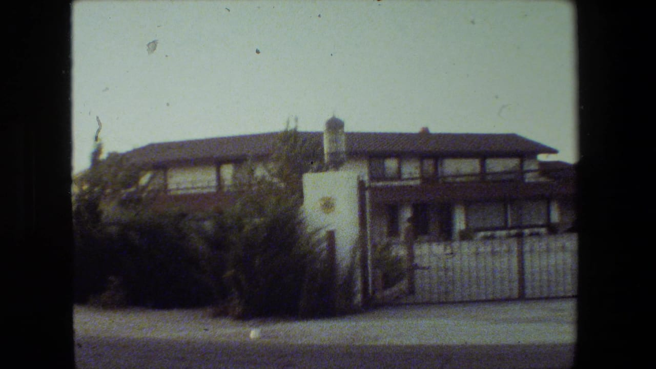 1981 Malibu 8mm frame showing a two-story stucco residence with dark roof trim, a white gate, shrubs, sidewalk, and street foreground.