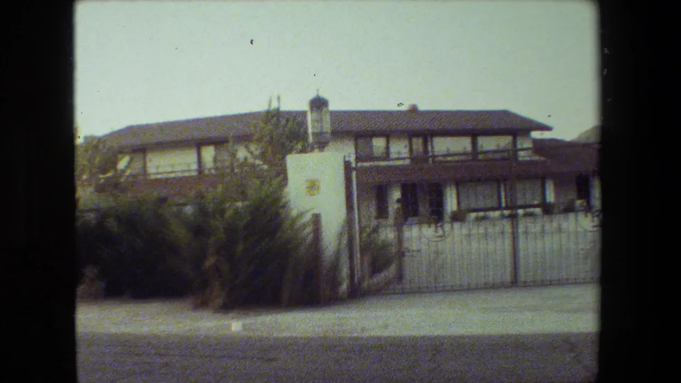 1981 Malibu home-movie frame of a gated two-story house with dark wood trim, white stucco, ornamental pillar lantern, bushes, and paved street.