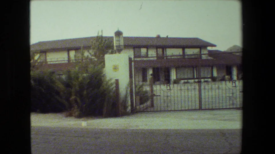 1981 Malibu 8mm frame centered on the gated residence, showing the white gate, stucco pillar, dark roofline, shrubs, sidewalk, and street.