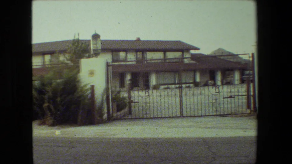 Final 1981 Malibu frame showing the two-story stucco house, white gated frontage, dark trim, pillar lantern, shrubs, sidewalk, and paved road.