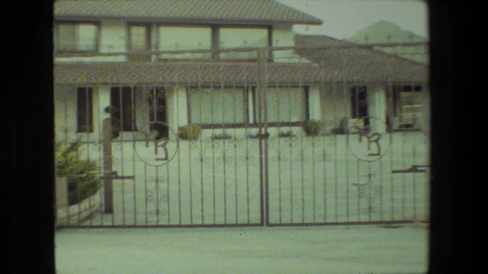 1981 Malibu-area frame with the front of a stucco house, tiled roof, windows, shrubs, and a metal gate filling the foreground.