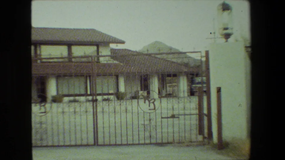 1981 Malibu-area home-movie frame showing a stucco residence behind a metal gate, with a decorative lantern on a pillar at the right edge.